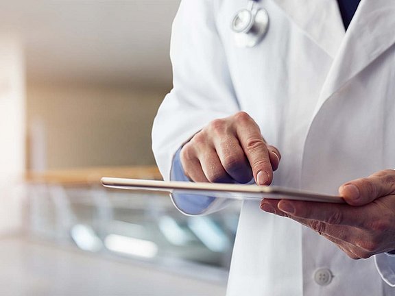 Ein Arzt im weißen Kittel benutzt ein Tablet in einer hellen, modernen Praxis. Um den Hals des Arztes ist ein Stethoskop zu sehen. Der Hintergrund ist verschwommen und lässt auf eine professionelle Innenumgebung schließen.