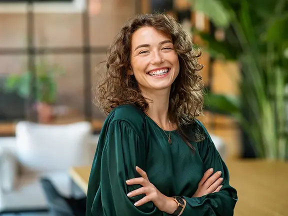 Eine lächelnde Person mit lockigem Haar und grüner Bluse steht mit verschränkten Armen in einer hellen Innenumgebung mit Pflanzen und unscharfem Hintergrund.